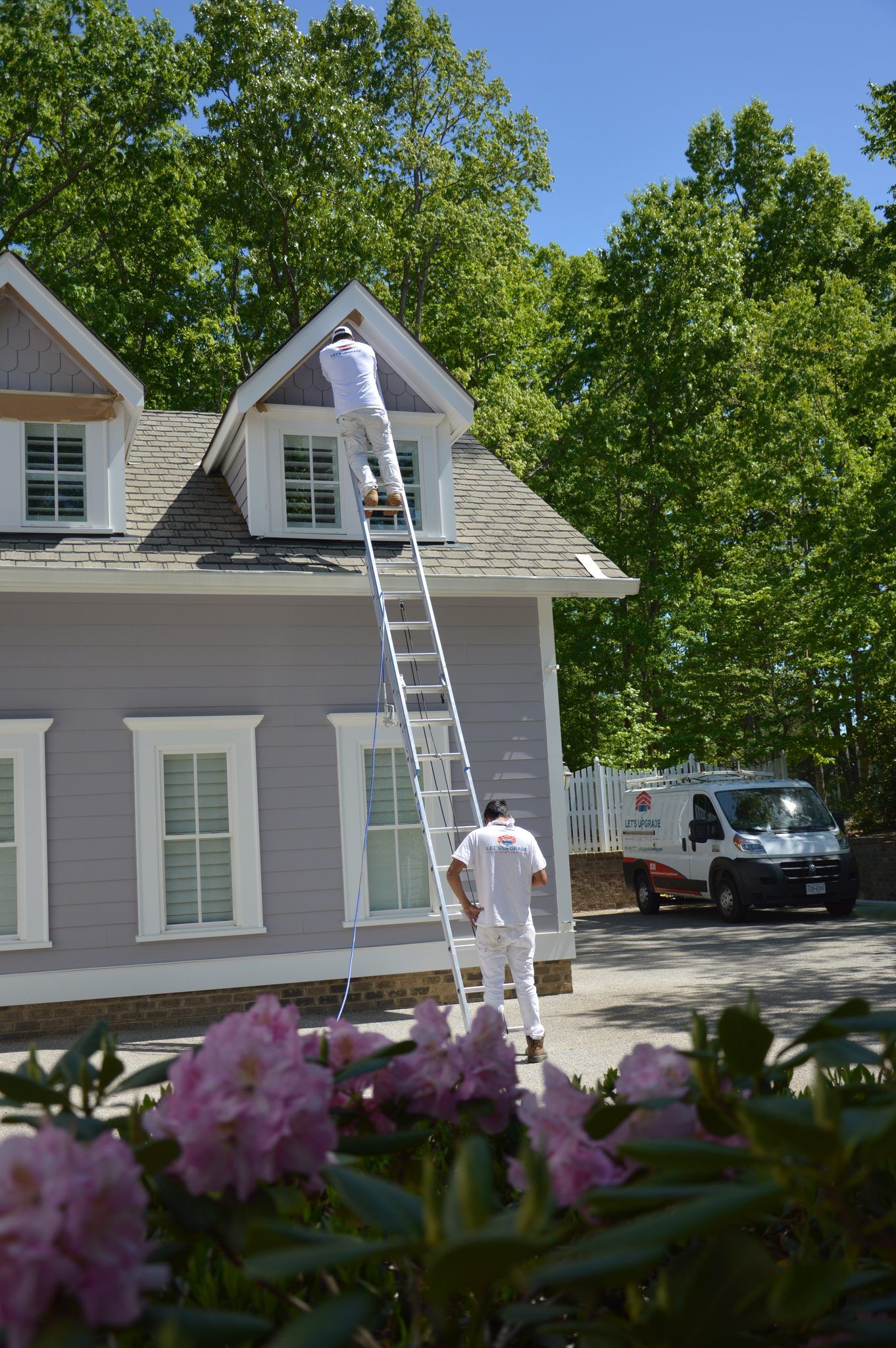 Painter On A Ladder Painting The Trim Of A Gray Two Story Home