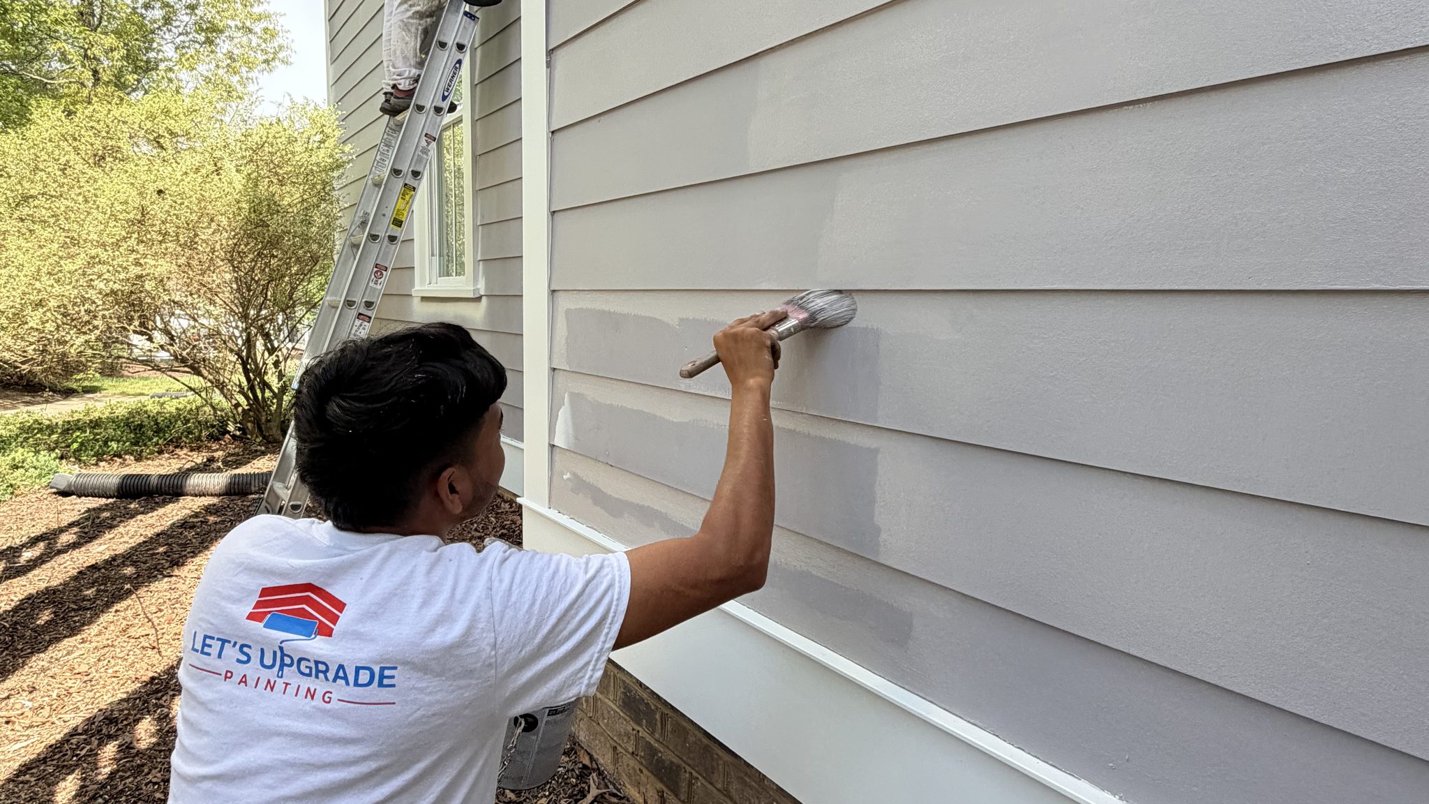 Painter Brushing Lap Siding On A Lavender Home